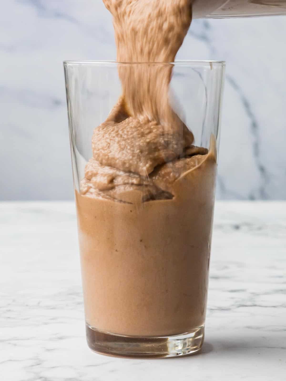 Pouring Nutella and banana smoothie into a glass from a blender, with a white marbled background.