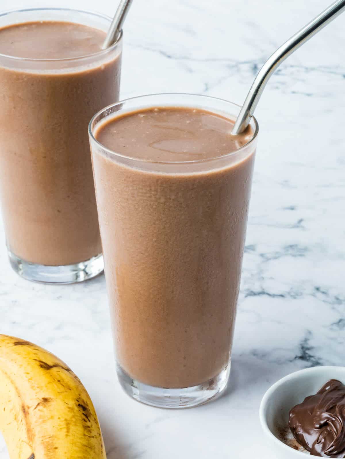 Two glasses of cold banana and Nutella smoothie with a banana and a small bowl of Nutella.
