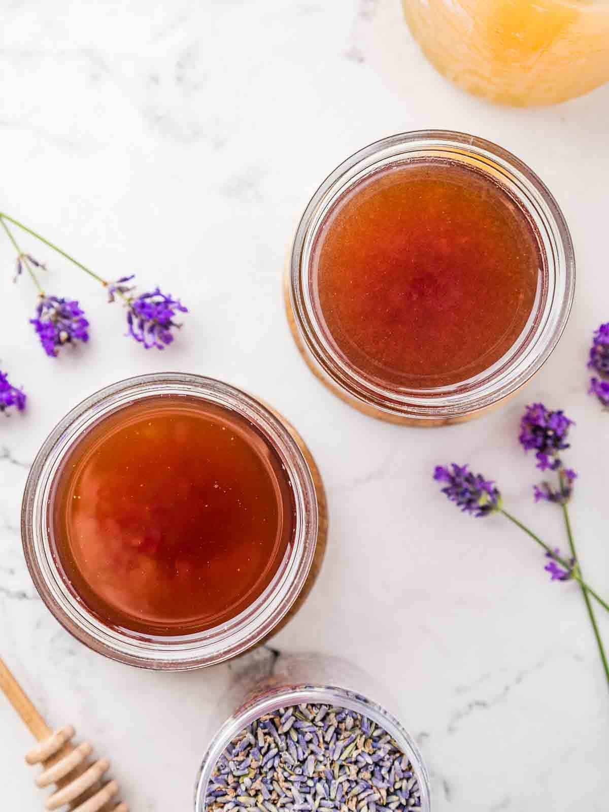 Two jars of lavender honey simple syrup with more lavender flowers around and a honey dripper.