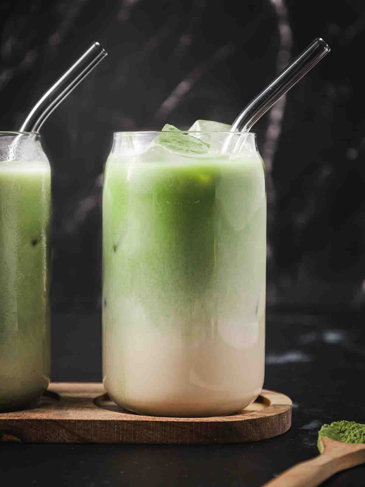 Two glasses of vanilla green tea latte with matcha powder around in a spoon.