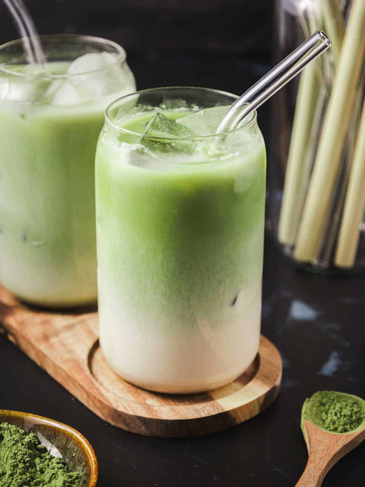 Two glasses of iced vanilla matcha latte served on a wooden plank, with straws and matcha powder around.