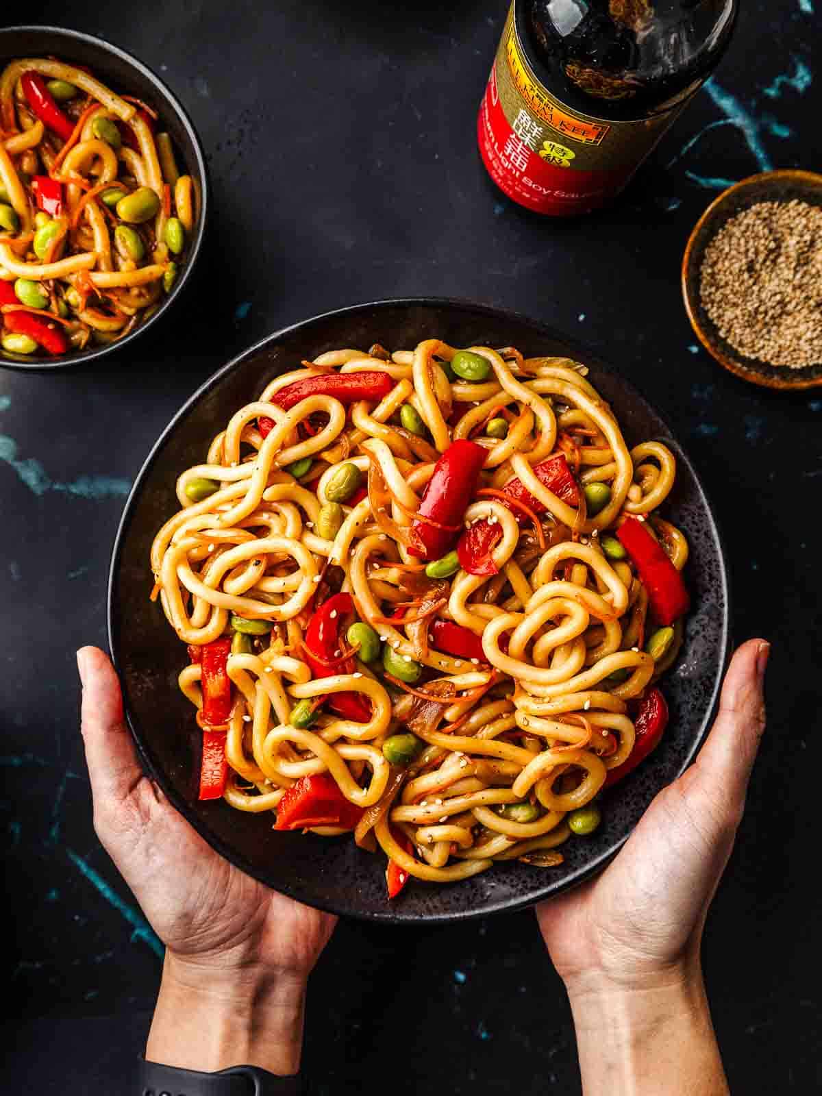 A plate of vegetable teriyaki udon noodles with sesame seeds sprinkled on top, hold with 2 hands.