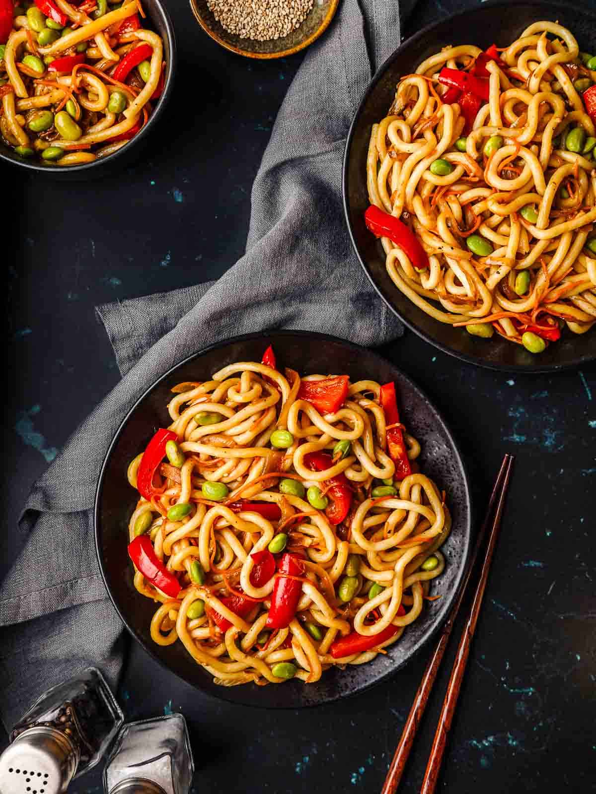 Three portions of teriyaki stir fry udon noodles on a black marbled background, with a grey napkin.