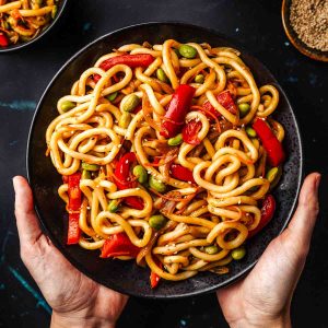 Vegetable teriyaki udon noodles with two hands holding the plate.
