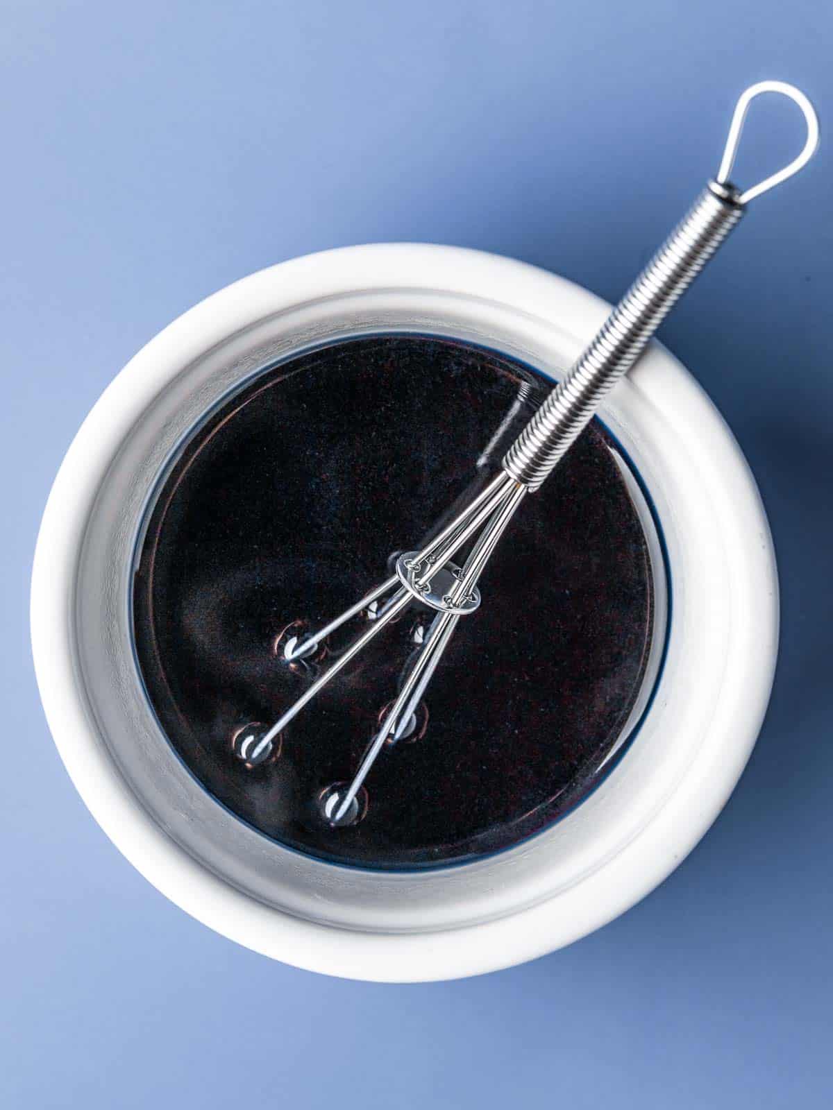 A bowl of butterfly pea flower tea, with a whisk, on a blue background.